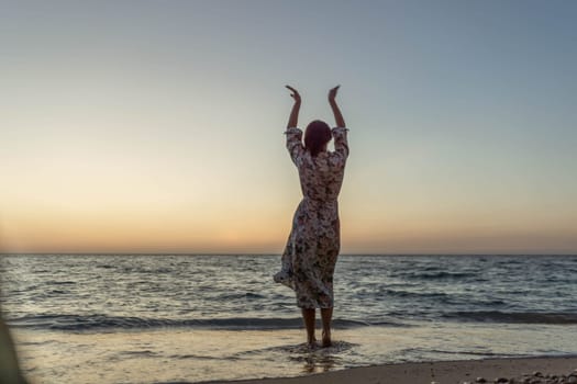 A woman in a dress stands on a beach with her arms raised, facing the ocean at sunset.