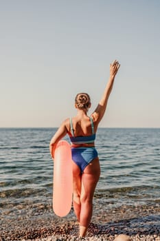 A woman is standing on the beach holding a pink inflatable tube. She is wearing a blue bikini and has her arms raised in the air. Scene is joyful and carefree