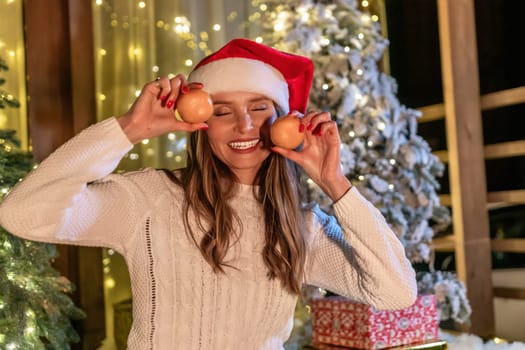A woman in a Santa hat playfully holds two Christmas ornaments over her eyes in front of a decorated Christmas tree.