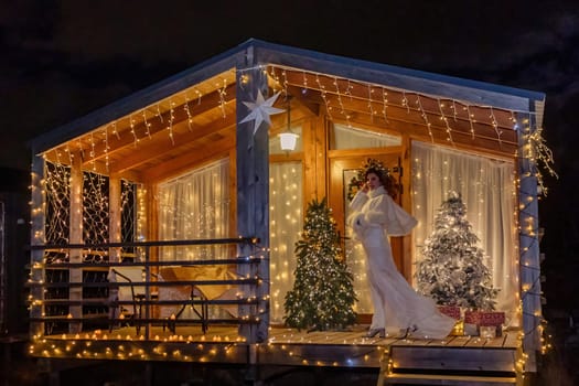 A woman in a white dress stands on the balcony of a small wooden cabin adorned with Christmas lights and two Christmas trees.