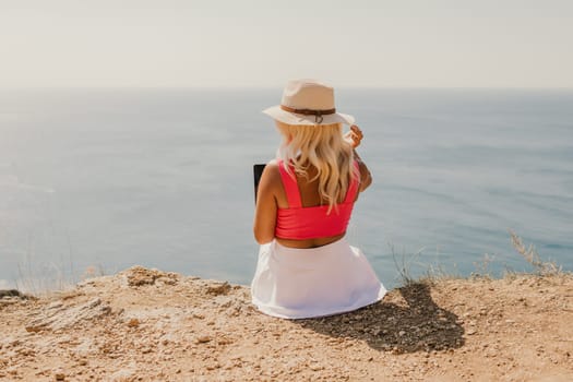 A woman is sitting on a rocky beach with a laptop open in front of her. She is wearing a pink top and a straw hat. Concept of relaxation and leisure