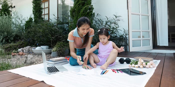 A mother and daughter spending quality time together outdoors, surrounded by art supplies, books, and a laptop on a sunny day.
