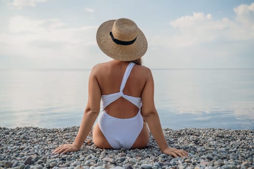 A woman wearing a white bikini and a straw hat is sitting on a beach. The beach is rocky and the sky is cloudy