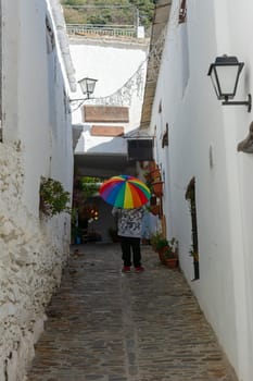 Person holding a rainbow umbrella walking down a narrow cobblestone alley with white walls and potted plants, Alpujarras, granada, andalucia, espa a,
