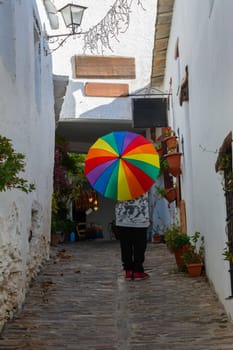 Close-up of a person holding a rainbow umbrella walking down a narrow cobblestone alley with white walls, Alpujarras, granada, andalucia, espa a,