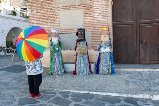 Person holding a rainbow umbrella standing in front of three statues outside a brick wall and wooden door, Alpujarras, granada, andalucia, espa a,