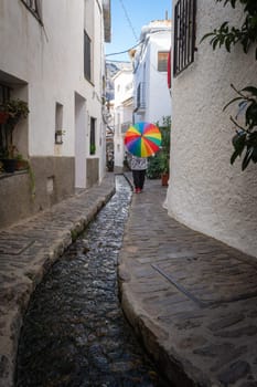 Person with a colorful umbrella walking along a narrow stone path with a stream in a white building alley, Alpujarras, granada, andalucia, espa a,