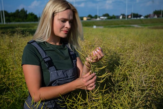 A female farmer in overalls inspects a rapeseed plant in a field, checking the ripening pods. The background features a rural landscape with slightly blurred houses and infrastructure under a partly cloudy sky.