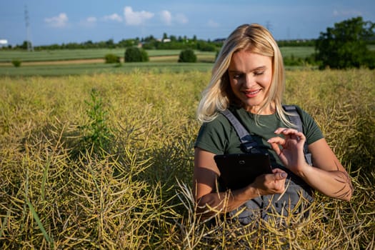 A woman farmer is checking the information about rapeseed using a tablet. She is gently touching the screen and checking if everything is ok with her harvest, what are the prices of rapeseed purchase. She is checking the information about her harvest. Farmer is also photographing the rapeseed with tablet camera. She owns a rapeseed field and is checking this year's harvest. She has blonde hair and smile showing her white teeth. The rapeseed is yellow and ready for harvest. Satisfied with the bountiful and lavish harvest, the woman farmer, adorned in working clothes and with tanned skin, gazes upon the expansive yellow landscape with a sense of fulfillment. With the blue sky adorned with fluffy white clouds overhead, she is standing forward. The day is warm and sunny.