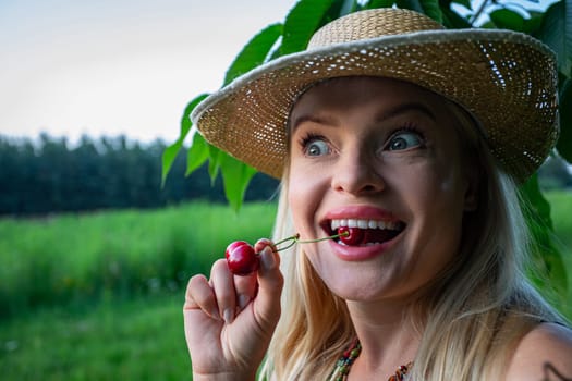 In the close-up shot, the woman's face and hands take center stage, her lips painted and full, complementing her vibrant blue eyes and playful smile. With an air of contentment, she eagerly anticipates indulging in the ripe cherries. In the background, a cherry tree branch and a blurred forest add to the serene atmosphere. Holding a single red cherry between her teeth, the stem protruding, she reveals her white teeth and tongue. The cherries, vibrant red and fully ripe, promise a burst of flavor. Completing her ensemble is a straw hat atop her head, perfect for the pleasant summer day. Her carefree smile and playful gaze add a sense of whimsy to the scene, as she looks off to the side with amusement. In this moment of simple joy, she embodies the essence of summertime bliss, basking in the warmth of the sun and the delight of nature's bounty.