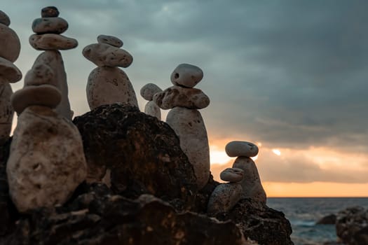 A group of rock cairns stand on a rocky outcrop overlooking the ocean at sunset. The sky is a dramatic mix of grey and orange.