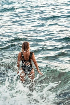A woman in a black swimsuit walks through the crashing waves of the ocean.