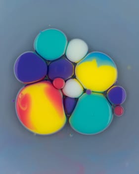 Multiple vibrant droplets of various colors blend and interact on a soft surface, creating an abstract and visually striking composition. The scene is illuminated by natural light.