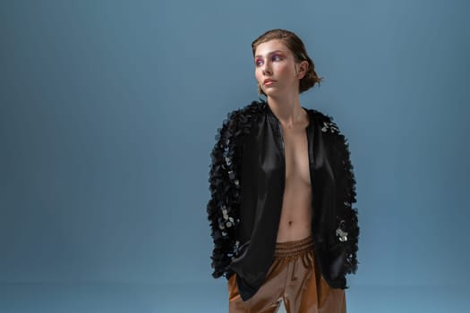 Sensual young female model with short hair in black fashionable jacket. Sexy woman with makeup in trendy outfit on naked body looking away against blue background.