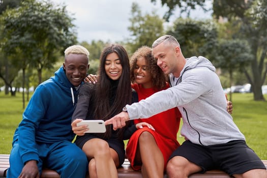 Young people of different nationalities taking selfie using smartphone sitting on bench in park in summer. Happy friends in sportswear spending leisure time together.