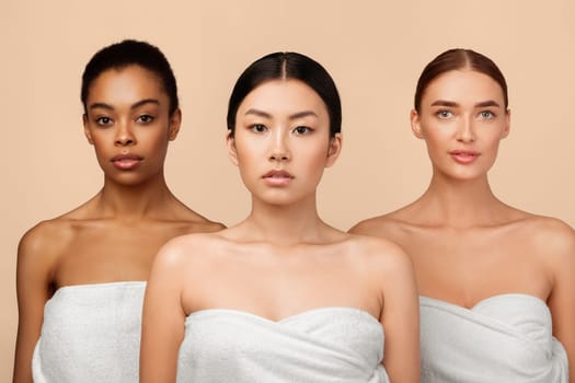 Portrait Of Three Sensual Diverse Girls Wrapped In Bath Towels Standing Posing In Studio Over Gray Background. Beauty Spa Concept.