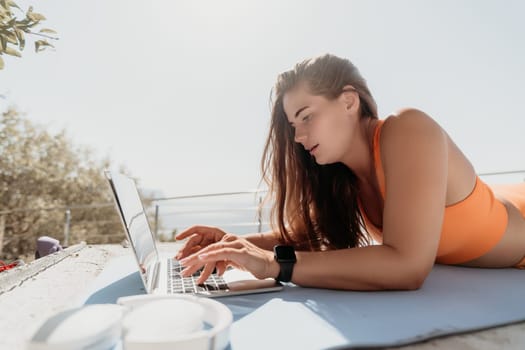 A woman wearing an orange swimsuit lays on a blue yoga mat, using her laptop outdoors.