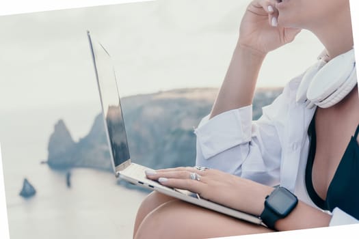 Digital nomad, woman in the hat, a business woman with a laptop sits on the rocks by the sea during sunset, makes a business transaction online from a distance. Freelance, remote work on vacation.