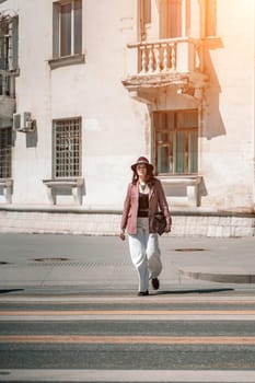 Woman city road crossing. Stylish woman in a hat crosses the road at a pedestrian crossing in the city. Dressed in white trousers and a jacket with a bag in her hands