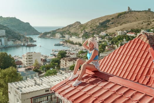 Woman sits on rooftop, enjoys town view and sea mountains. Peaceful rooftop relaxation. Below her, there is a town with several boats visible in the water. Rooftop vantage point