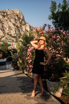 A woman in a black dress and straw hat stands barefoot in front of a flowering bush, with a large mountain in the background. family vacation concept
