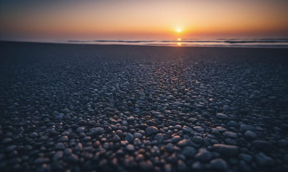 A low angle shot of a beach covered in small, smooth pebbles. The sun is setting in the distance, casting a golden light on the water