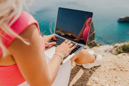 A woman is sitting on a rock by the ocean, typing on her laptop. Concept of relaxation and leisure, as the woman enjoys the beautiful view of the water while working on her computer