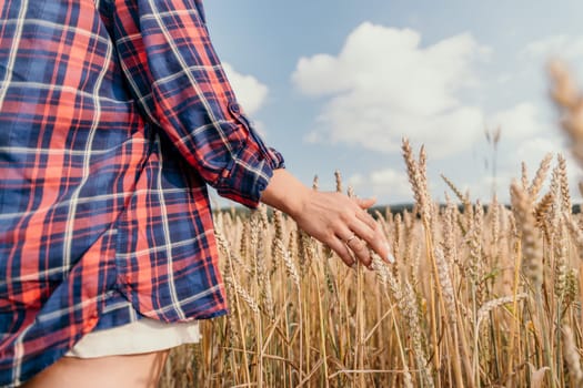 A woman wearing a red and blue plaid shirt reaches out to touch stalks of wheat in a field.
