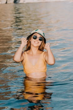 A woman in a yellow bikini and a bucket hat stands in a pool, smiling and adjusting her sunglasses.