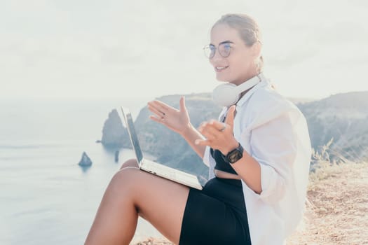 Digital nomad, woman in the hat, a business woman with a laptop sits on the rocks by the sea during sunset, makes a business transaction online from a distance. Freelance, remote work on vacation.