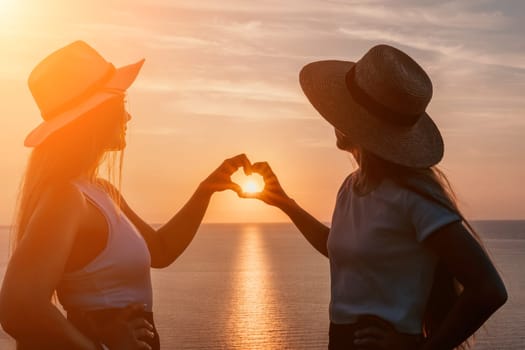Two women in silhouette at a sunset make a heart shape with their hands over a body of water.