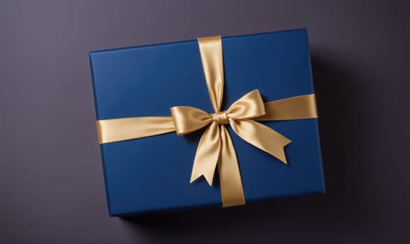 A square, blue gift box is wrapped in gold ribbon. The ribbon is tied into a bow in the center of the box. The box is sitting on a dark gray surface.