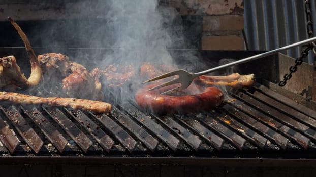 Video showing various meats being grilled on a barbecue, using a large fork to flip and check the doneness of the steaks over open flames and hot coals.