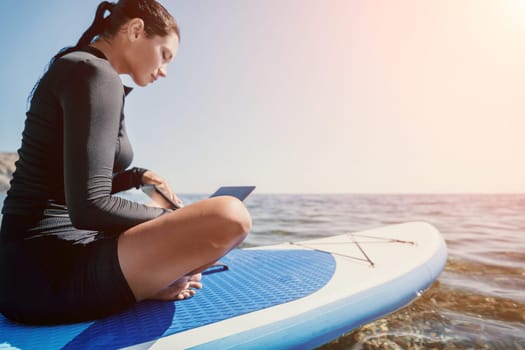 Digital nomad, woman in the hat, a business woman with a laptop sits on the rocks by the sea during sunset, makes a business transaction online from a distance. Freelance, remote work on vacation.
