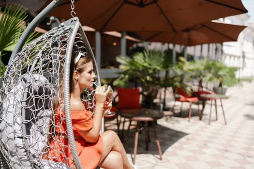 A woman in an orange dress is sitting in a chair with a drink in front of her. She is looking at the camera and she is enjoying her drink. The scene is set in a patio or outdoor area