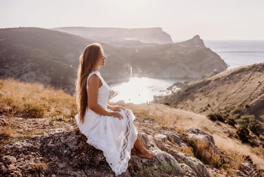 A woman in a white dress sits on a rock overlooking a body of water. The scene is serene and peaceful, with the woman enjoying the view and the calmness of the surroundings