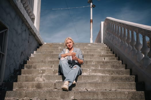 A woman in a suit and jeans sits on a set of stairs. She is holding a cup and she is smiling