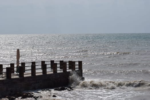 big sea waves splashing against a pier. The pier cuts the sea waves off the coast during a stormy sunny day.