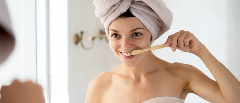A young girl brushes her teeth near the mirror after relaxing in the bathroom, the woman is wrapped in a towel and takes care of the health and beauty of her body and teeth, gums.