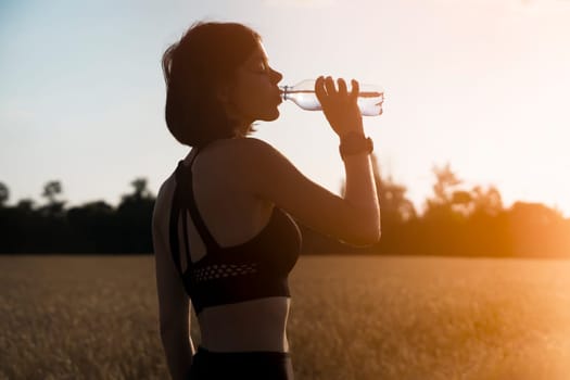 A young girl takes a break during an intense workout and jogging and drinks refreshing cool water from a bottle, a woman trains outdoor in the evening at sunset.