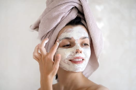 A young girl in a towel on her head and body stands near the mirror in the bathroom and applies a clay mask to her face, the woman takes care of her health and beauty.