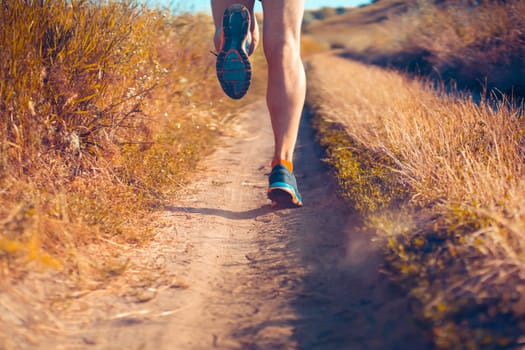 Athlete runner legs running on a close-up of a trail across a field on a shoe. Fitness and warm-up, young man with sun effect in the background.