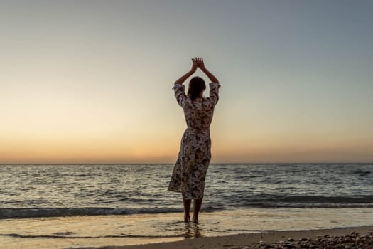 A woman in a dress stands on a beach facing the ocean at sunset, her arms raised above her head.