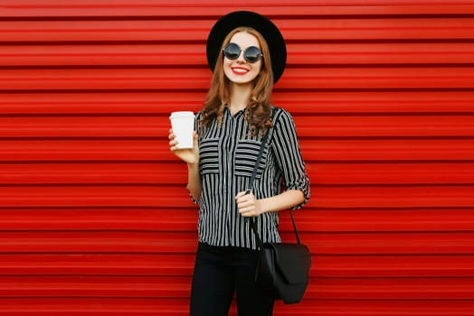 Stylish young woman with coffee cup wearing a black white striped shirt, round hat and handbag clutch on city street over red wall background