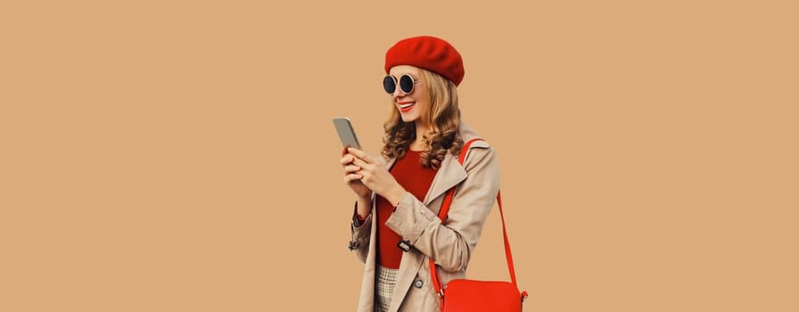 Autumn portrait of stylish happy smiling young woman with phone, beautiful girl holding smartphone in red french beret hat on brown background