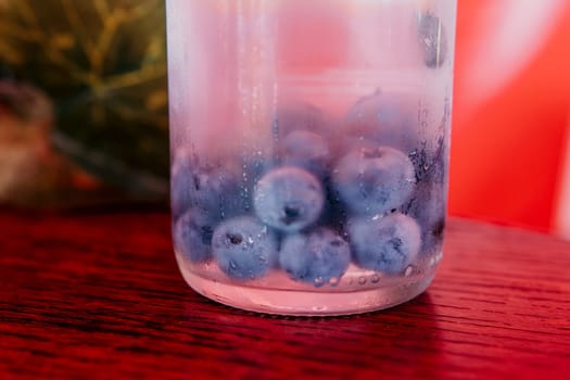 A glass filled with blueberries is sitting on a red wooden surface. The condensation on the outside of the glass makes the blueberries appear even more vibrant.
