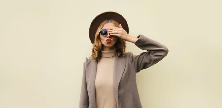 Autumn portrait of beautiful young woman blowing a kiss in round hat, stylish girl posing on brown background