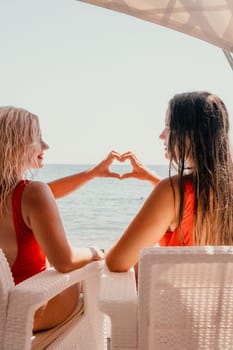 Two women in red swimsuits are seated on white wicker chairs, creating a heart shape with their hands. The ocean is visible in the background, with a sunny atmosphere.