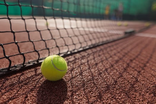 A close-up of a tennis ball resting on a clay court, with a net in the foreground and blurred players in the background