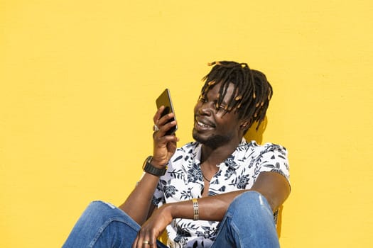 smiling young black man sitting against a yellow wall recording a voice message with his smartphone, technology and lifestyle concept, copy space for text